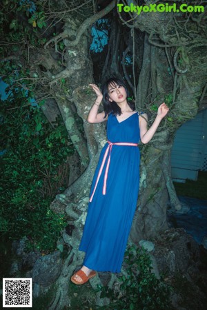 A woman in a blue dress standing next to a tree.