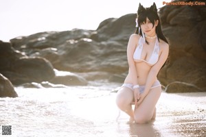 A woman in a white bikini sitting on a rock by the water.